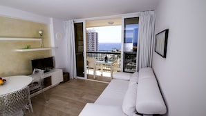 A modern living room with a white sofa, a dining table with chairs, a TV, and a view of the sea through large glass doors. - 150 m from the sandy beach, fantastic sunsets, WiFi, satellite TV, swimming pools (Costa Adeje)