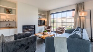 Una sala de estar moderna con un sofá, sillas, una mesa de centro y un televisor. Hay estanterías con objetos decorativos y una ventana con cortinas.