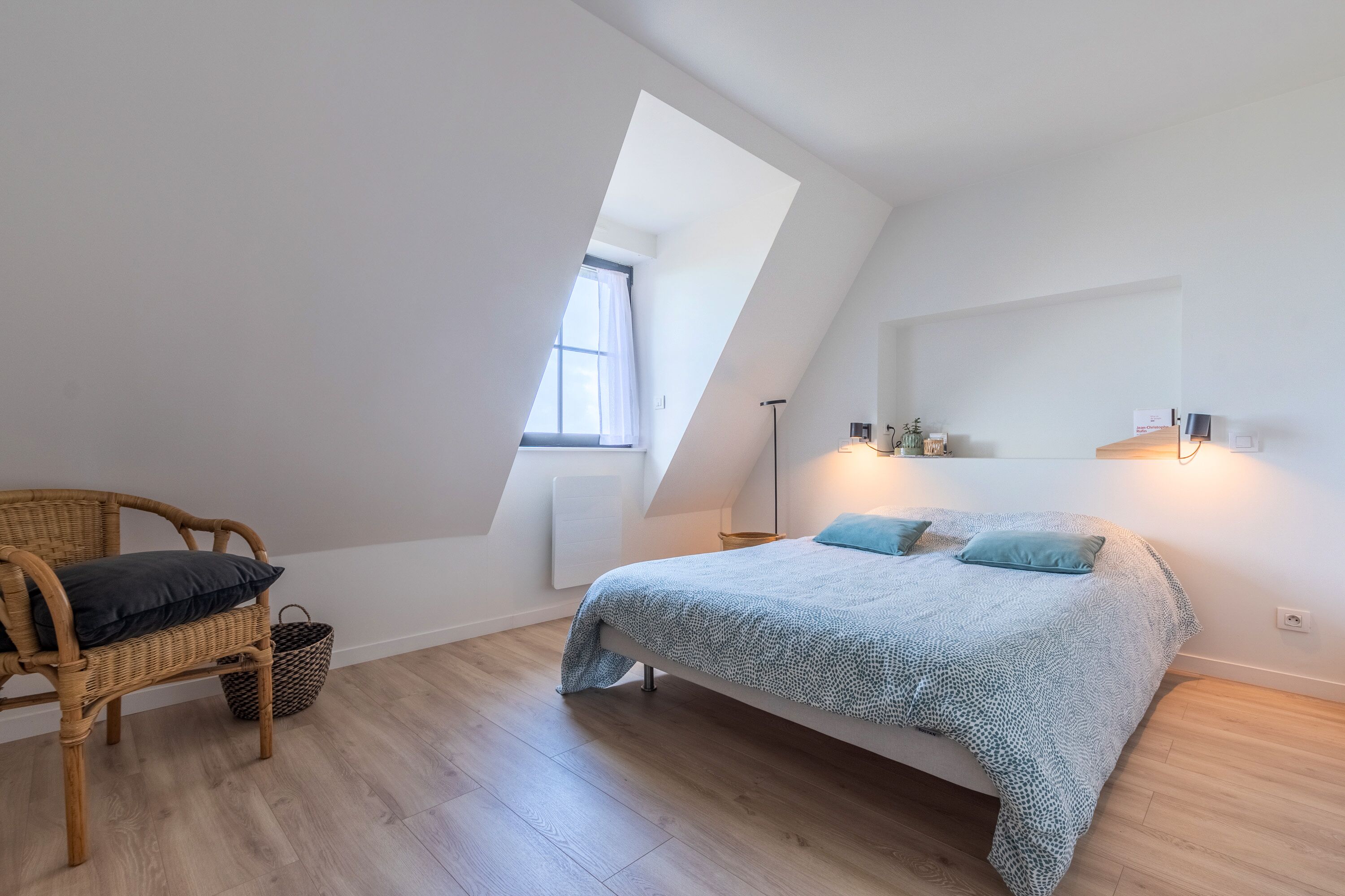 Een slaapkamer met schuin dak, een bed met beddengoed met een patroon, een rieten stoel en een nachtkastje met een lamp.