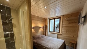 A bedroom with a wooden ceiling, a bed with a patterned bedspread, a window with a view, and a shower area with a tiled wall. - GENTIANES 1 – Lovely apartment in the village center and close to the slopes – Wi-Fi (La Clusaz)
