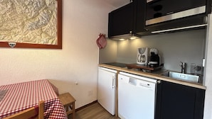 A compact kitchen with a dishwasher, a coffee maker, and a microwave. There is a small table with a checkered tablecloth and a chair. A framed map is hanging on the wall. - RESIDENCE 2-235 - Village-center studio, close to the slopes - Wi-Fi - Garage (La Clusaz)