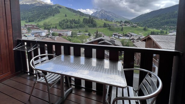 A balcony with a metal table and chairs, overlooking a mountainous landscape with buildings and greenery. - RESIDENCE 2-235 - Village-center studio, close to the slopes - Wi-Fi - Garage (La Clusaz)