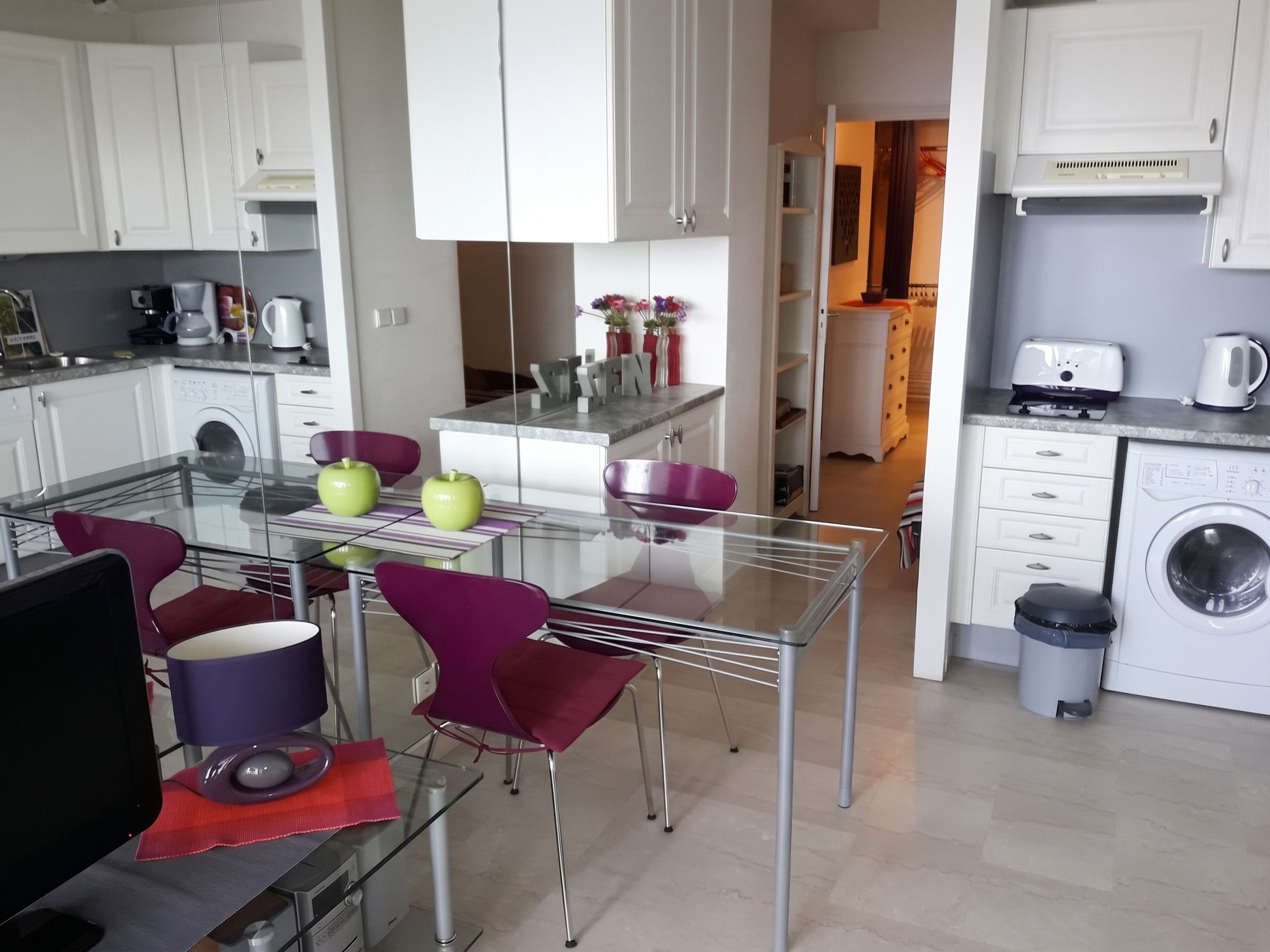 A modern kitchen with white cabinets, a glass dining table, and purple chairs.