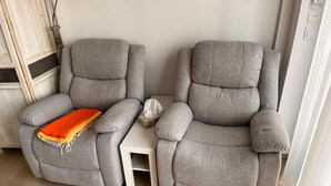 Zwei graue Liegesessel mit einem kleinen weißen Beistelltisch, einem Heizgerät und einem Fenster mit durchsichtigen Vorhängen.