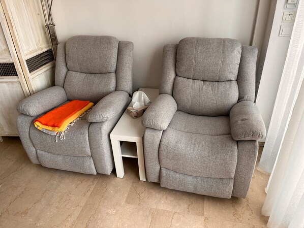 Two grey reclining chairs with a small white side table, a heater, and a window with sheer curtains.
