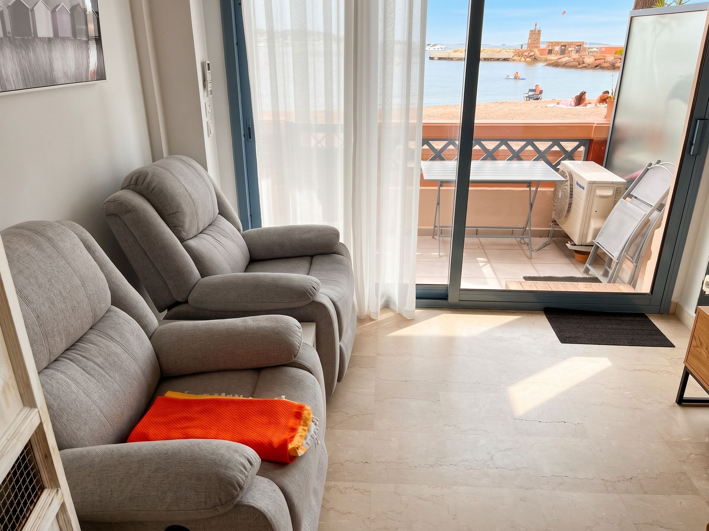 Een woonkamer met een bank, uitzicht op een strand en een balkon met een tafel en stoelen.