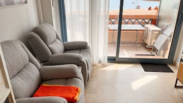 Een woonkamer met een bank, uitzicht op een strand en een balkon met een tafel en stoelen.