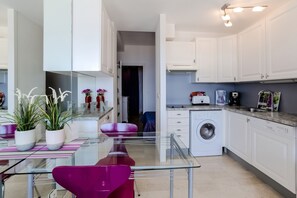A modern kitchen with white cabinets, a washing machine, and a glass dining table with a pink chair. - Homerez - Apartment in Théoule-sur-Mer. (Théoule-sur-Mer)