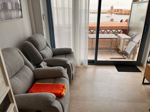A living room with a grey sofa, a small table, and a balcony with a view of the sea.