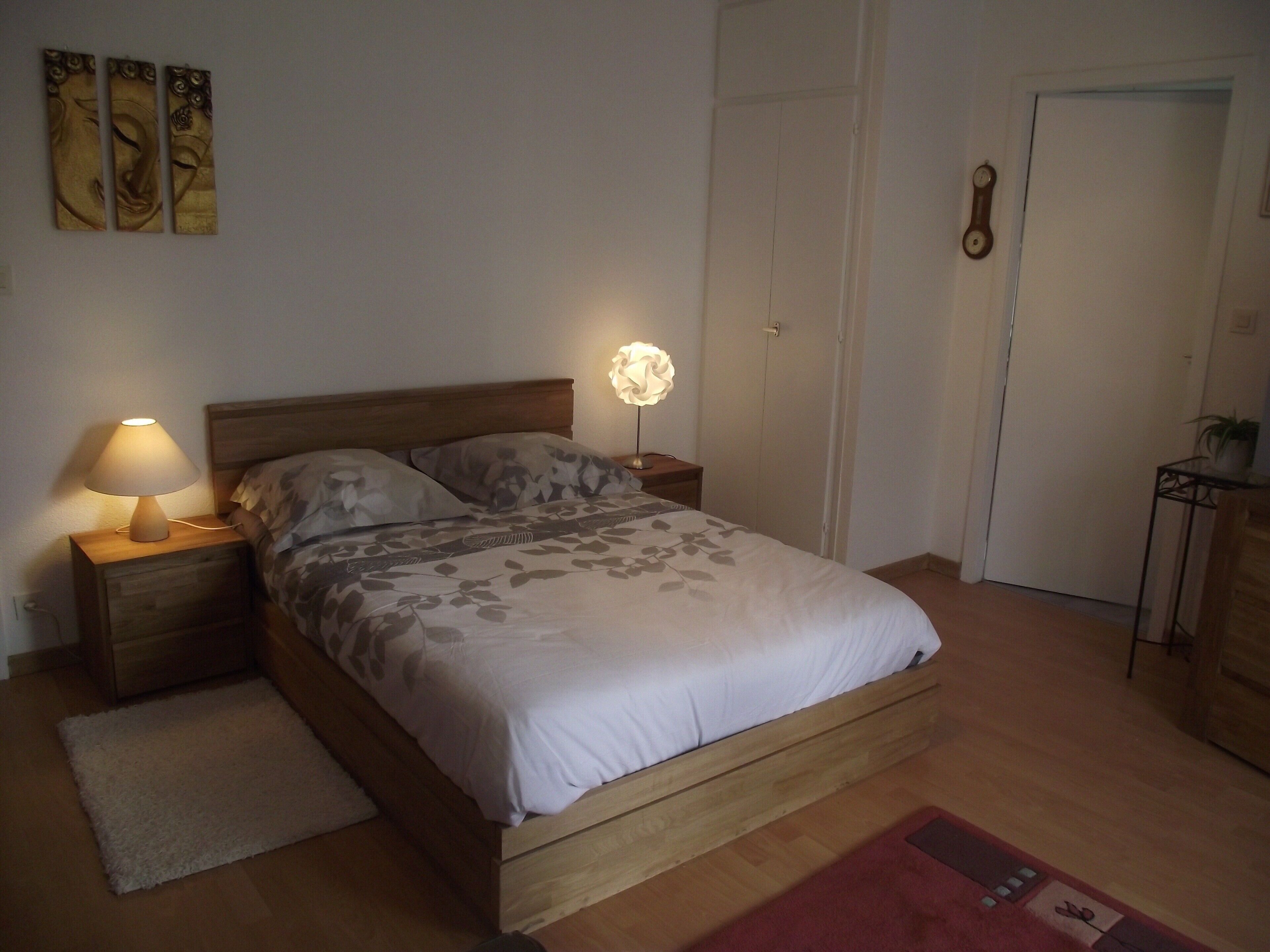 Une chambre à coucher avec un lit en bois, deux lampes de chevet, une table de chevet, une porte et une petite table sur laquelle est posée une plante.
