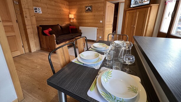 Interior em madeira com uma mesa de jantar posta para quatro pessoas, um sofá e uma televisão fixada na parede.
