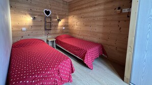 Um quarto pequeno e acolhedor, com paredes de madeira e duas camas individuais com roupa de cama vermelha. Há uma pequena mesa e uma prateleira fixada na parede com uma decoração em formato de coração.