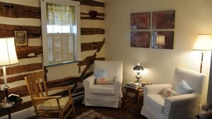 A cozy living room with a wooden wall, a staircase, two white armchairs, a wooden rocking chair, and a red rug.