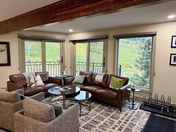 A living room with a brown leather sofa, a round coffee table, and a patterned rug. There are large windows with a view of greenery outside.