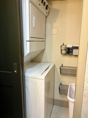 A laundry room with a stacked washer and dryer, a trash bin, and wire baskets on the wall.