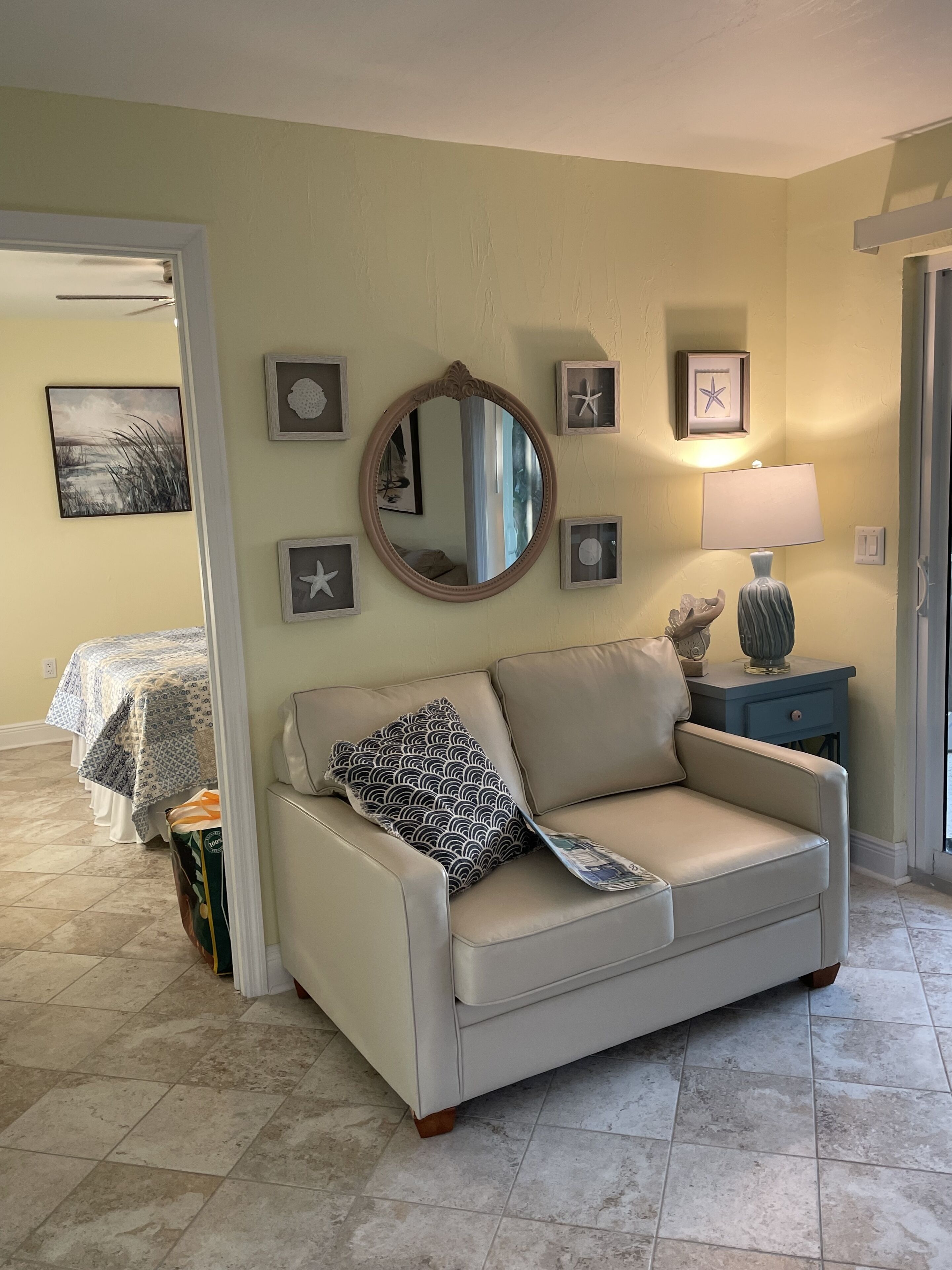 A living room with a beige sofa, a round mirror, framed artwork, and a lamp on a side table.