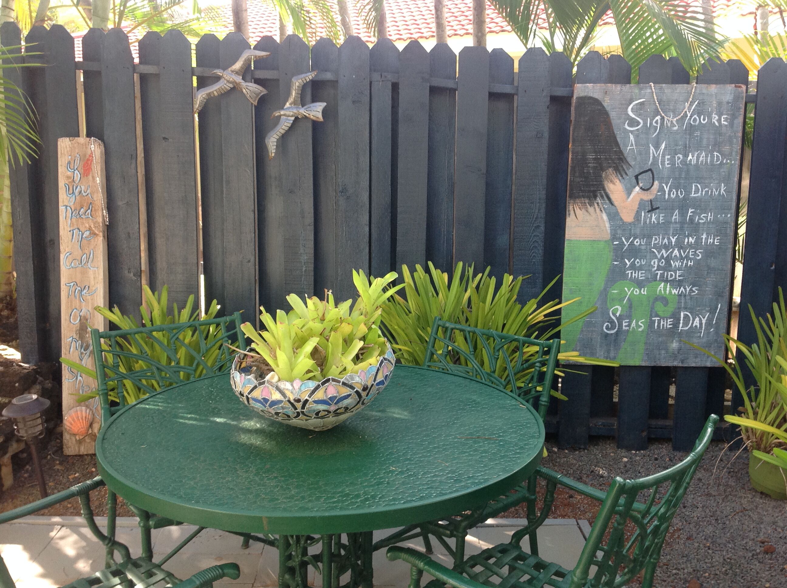 A round green table with a decorative bowl in the center, surrounded by green chairs. A wooden fence with bird decorations and a sign with text is in the background.