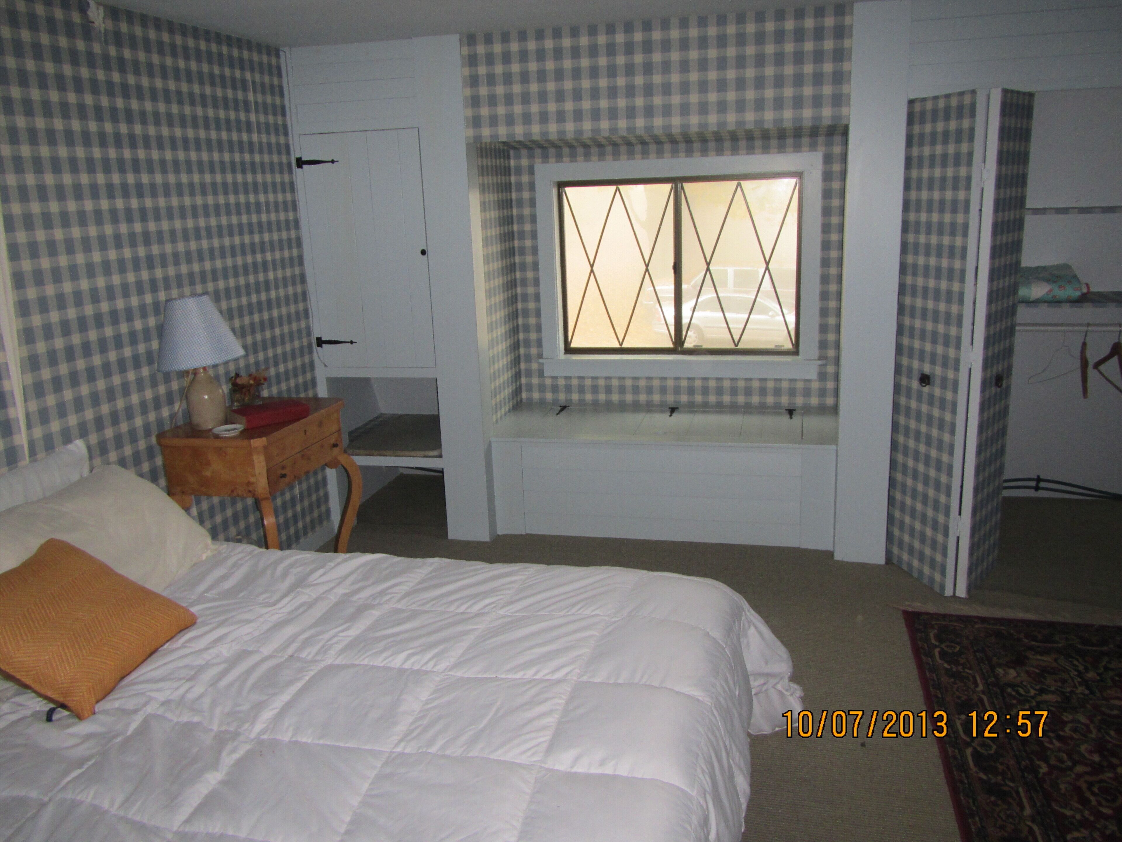 A hotel room with a bed, a bedside table with a lamp, a window with a checkered blind, and a door leading to another room.