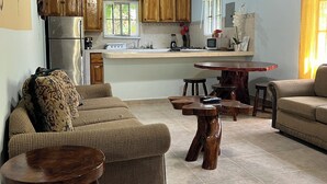 Un vaste espace de vie comprenant un canapé, une table basse et une table à manger avec des tabourets. On aperçoit, au fond, une cuisine avec des meubles en bois et un réfrigérateur.