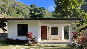 Un petit bâtiment blanc avec un toit en tuiles, une porte en bois et une fenêtre. On y voit deux chaises à l’extérieur et un pot de fleurs devant l’entrée.