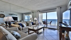 A spacious living room with a large sofa, a coffee table, and a TV. There is a balcony with a view of the sea.