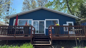 Une maison bleue avec une terrasse en bois, deux grandes fenêtres et un parasol rouge.