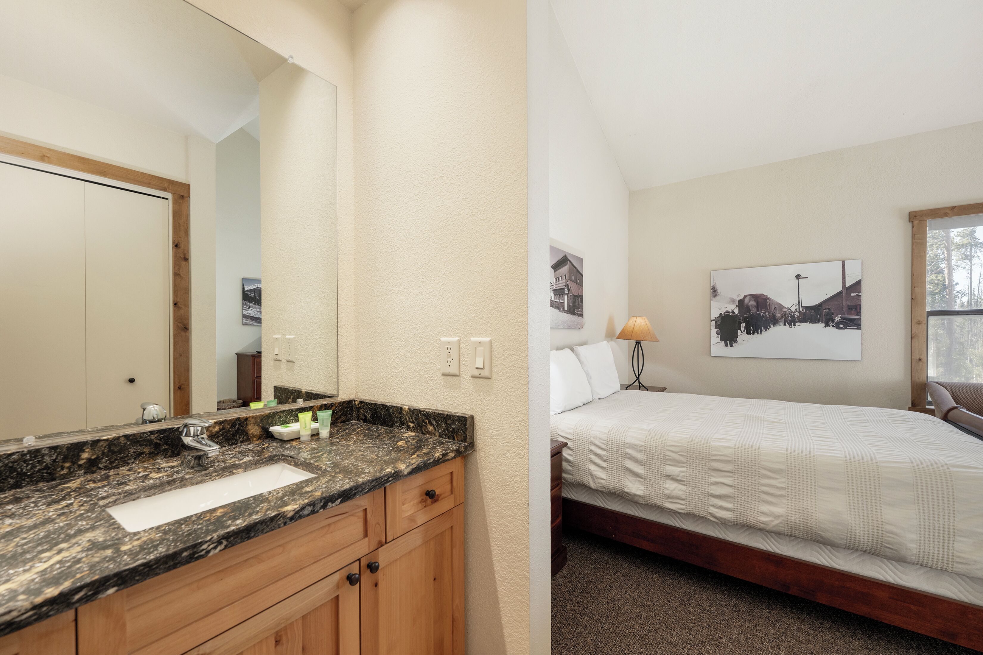 Un baño con lavamanos de encimera de granito, armarios de madera y un espejo grande. La habitación tiene una decoración en tonos neutros, con paredes color beige y cabecero de cama de madera.