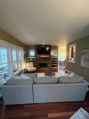 A living room with a sectional sofa, a fireplace, a TV mounted on a stone wall, and large windows. - Beautiful home in Picturesque setting just steps from Lake Michigan! (Fennville)