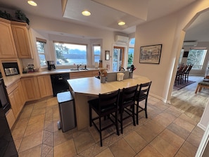 A kitchen with wooden cabinets, a built-in dishwasher, a large island with chairs, and a view of the outdoors through large windows.
