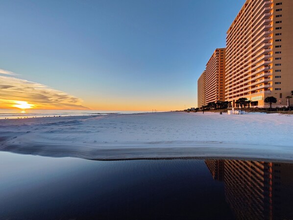 Un hotel frente a la playa con una piscina reflectante, vista despejada del océano y una puesta de sol al fondo.