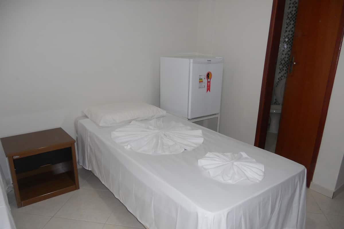 Quarto pequeno e limpo com uma cama de solteiro, mesa de cabeceira de madeira, frigorífico branco e porta para outro espaço.