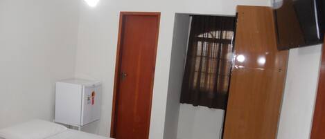 Una habitación con cama, un refrigerador blanco pequeño, una puerta de madera, una ventana con cortinas y un ventilador de techo.