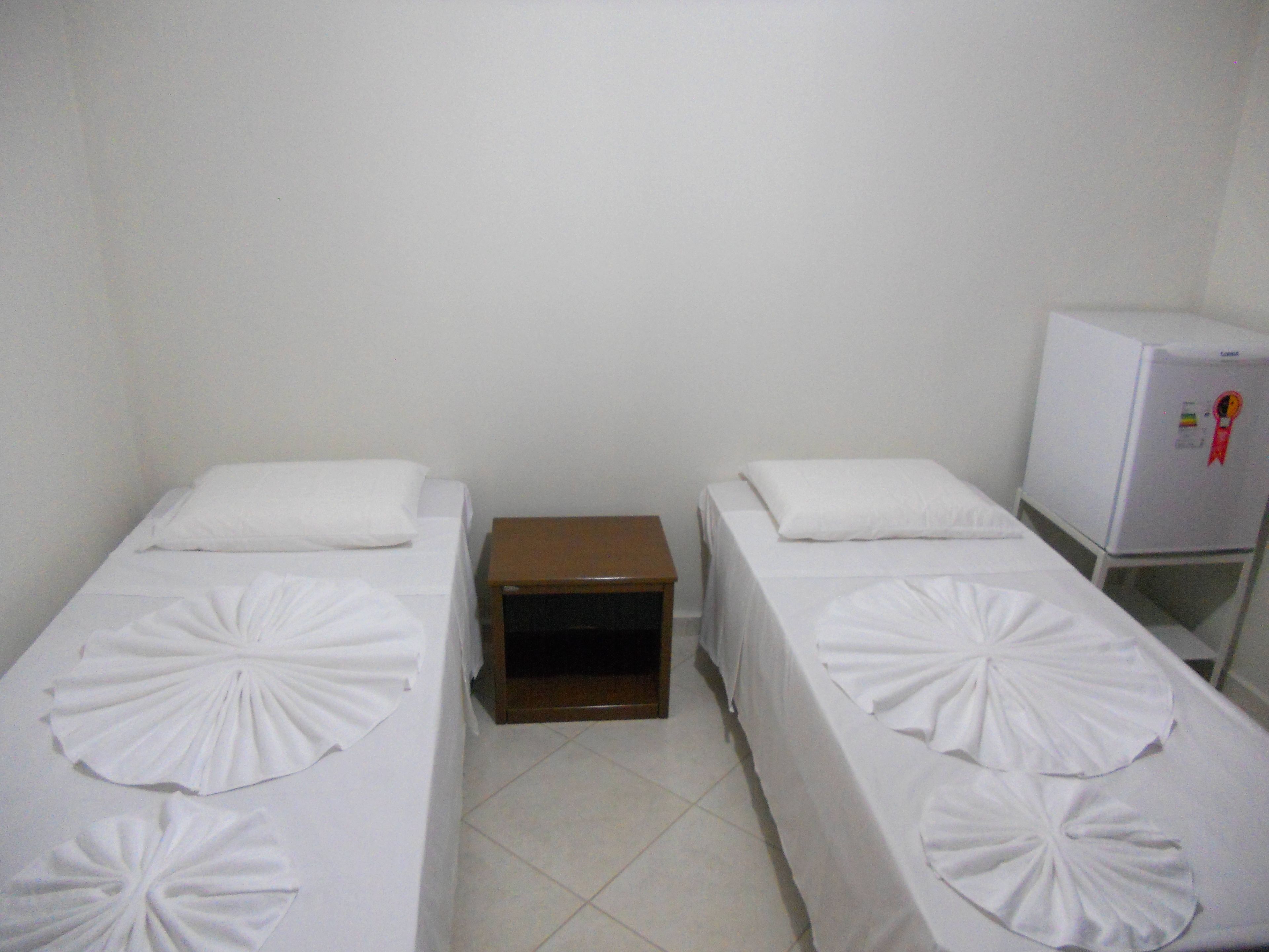 Two single beds with white linens and decorative pillows, a small wooden nightstand, and a white refrigerator.