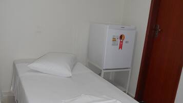 Habitación con cama individual, cubrecama blanco, almohada, mesita pequeña y refrigerador.