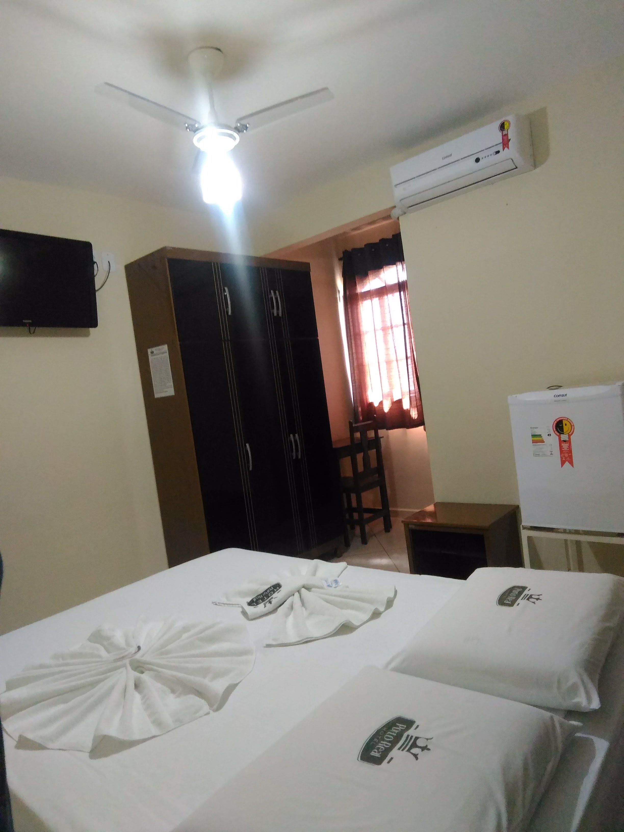 Una camera d'albergo con due letti, una ventola a soffitto, un climatizzatore, una televisione, una scrivania e una sedia.