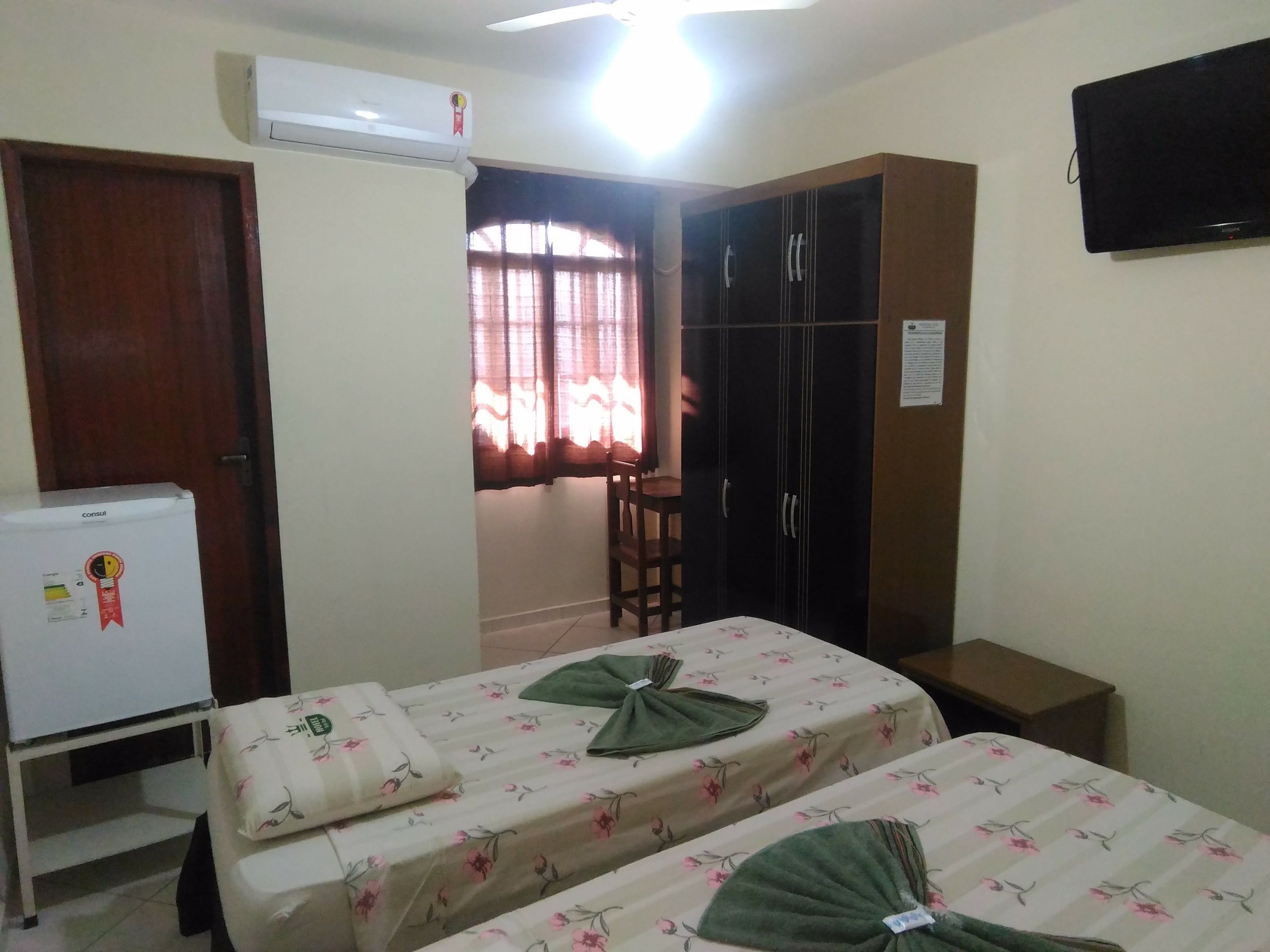 A room with two beds, a wardrobe, a desk, a chair, a television, and an air conditioning unit.