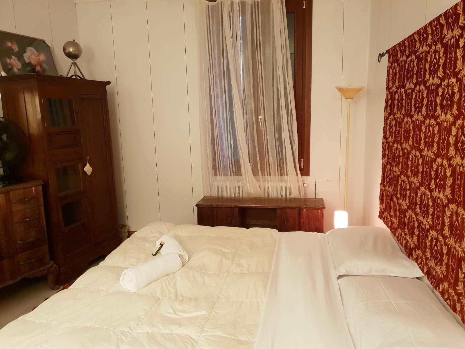 Ein Schlafzimmer mit einem Bett, einem Holzschrank, einem Nachttisch, einer Lampe und einem Fenster mit durchscheinendem Vorhang.