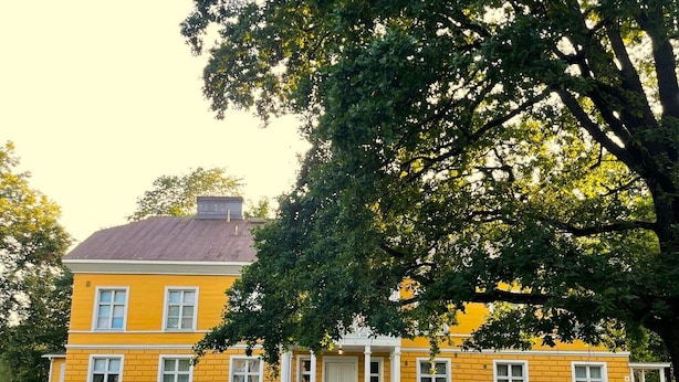 A two-story yellow building with a white door and windows, surrounded by a well-maintained lawn and large trees. - Tehtaan Hotelli (Karkkila)
