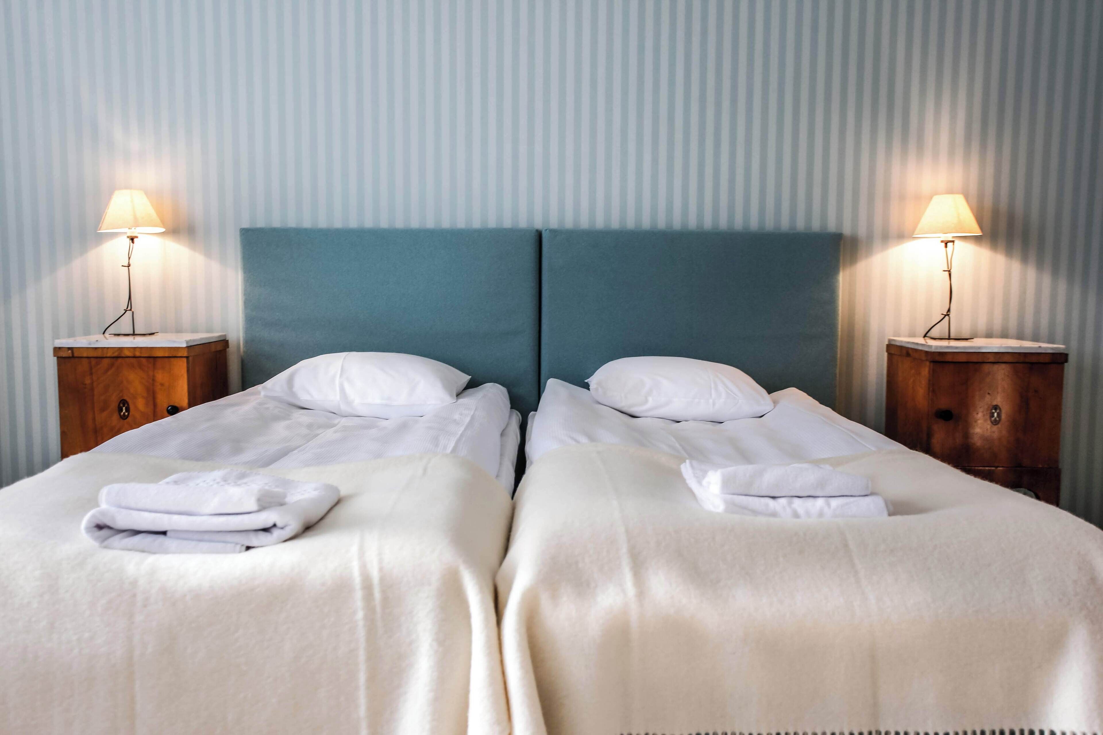 A hotel room with two beds, each with a white bedspread and pillows, flanked by wooden nightstands with lamps.