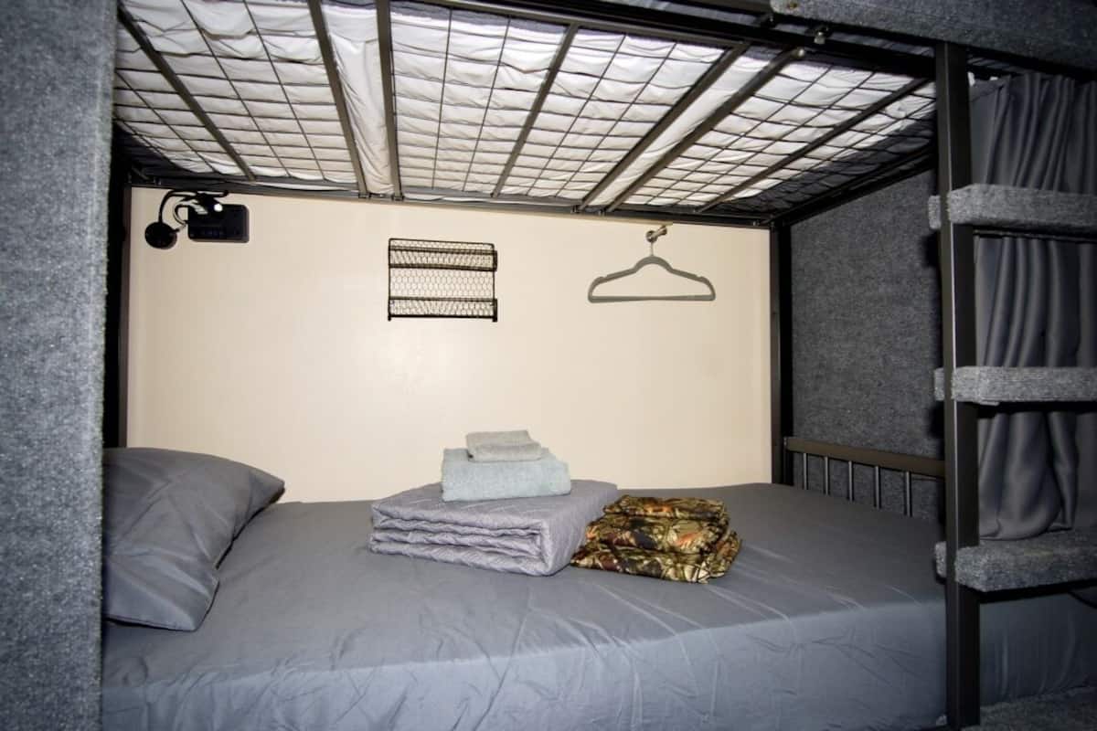 A bunk bed room with a metal frame, a headboard, a wall-mounted light, and a small shelf with a towel and a blanket.