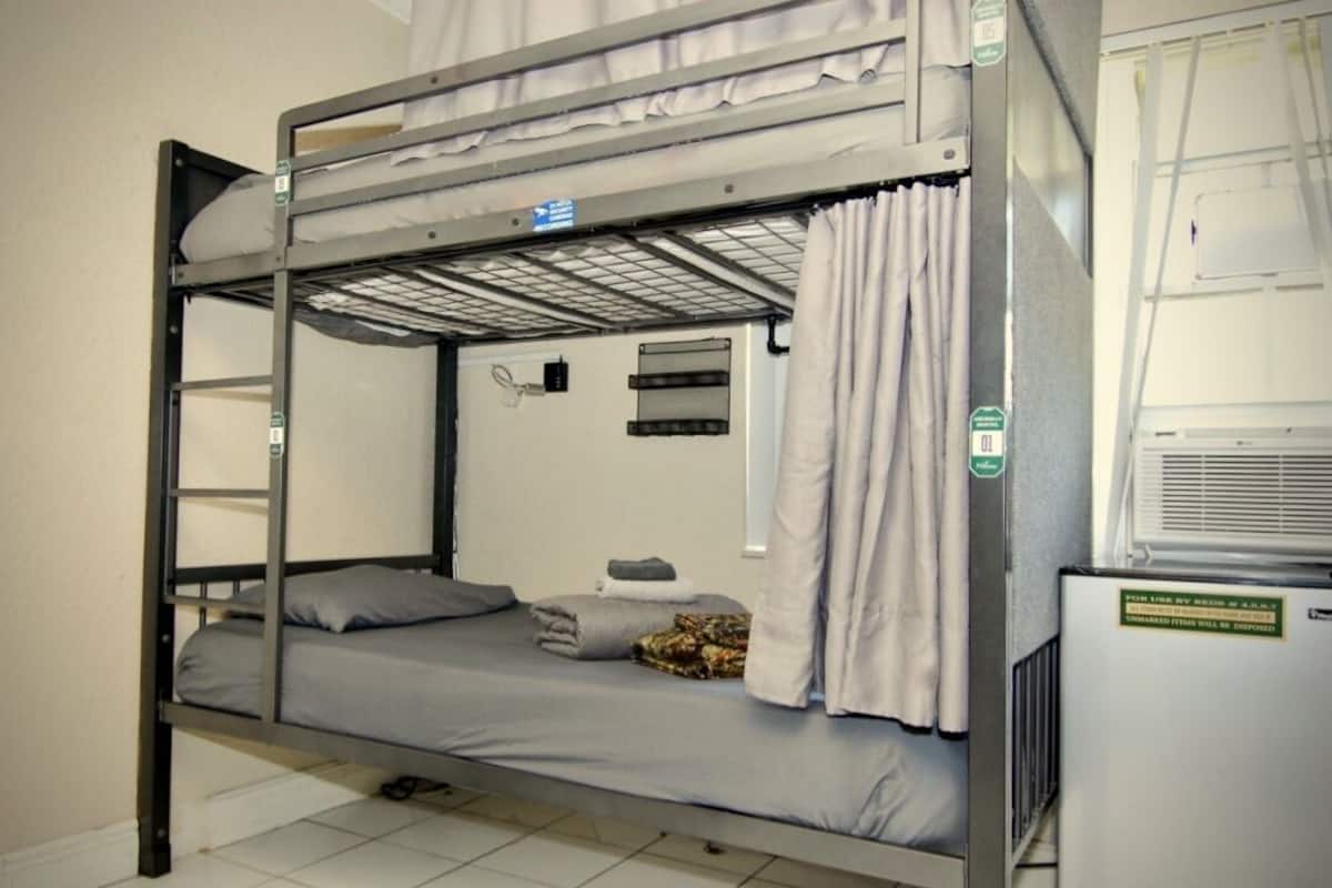 A bunk bed with a mattress, pillows, and a blanket. There is a window with curtains and a thermostat on the wall.