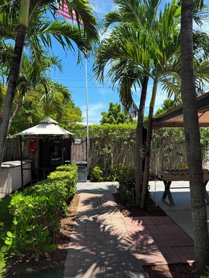A pathway leading to a covered area with a sign indicating an exit, surrounded by lush greenery and palm trees. - Sheridan Hostel (Dania Beach)