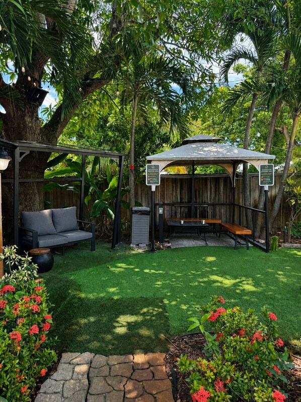 A covered outdoor seating area with a wooden table and benches, surrounded by lush greenery and a well-maintained lawn. - Sheridan Hostel (Dania Beach)