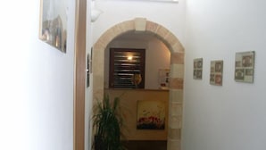 A hallway with a tiled floor, a potted plant, a framed artwork, and a stone archway leading to a room with a window and blinds. - B&B Li Cuti (Tiggiano)