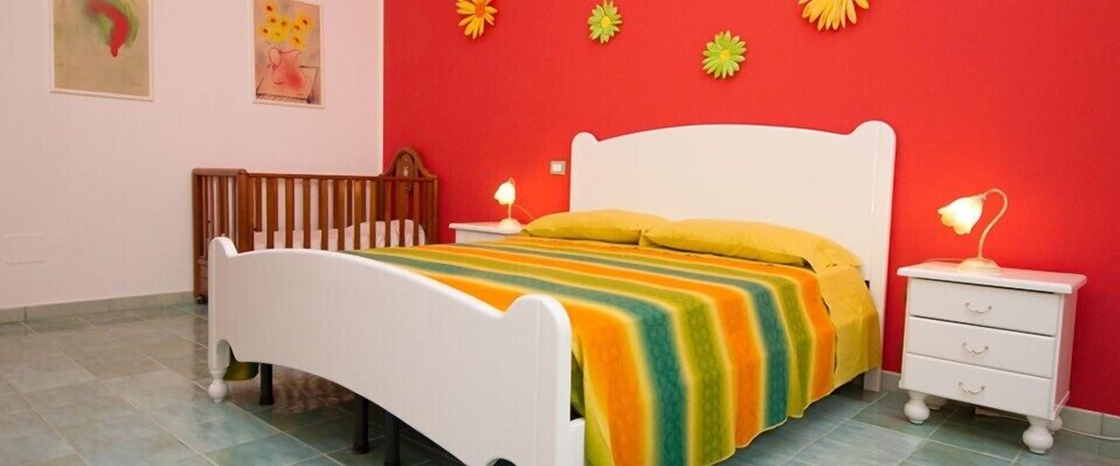 Un dormitorio con una cama blanca, una colcha rayada de colores, una cuna de madera y una mesita de noche blanca con una lámpara.