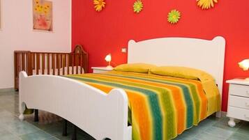 Un dormitorio con una cama blanca, una colcha rayada de colores, una cuna de madera y una mesita de noche blanca con una lámpara.