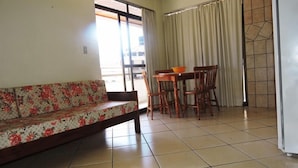 A living room with a sofa, a dining table with chairs, and a view through a window. - Residencial Baleia Franca (Florianópolis)