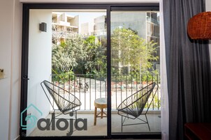 Un balcón con dos sillas de alambre negras y un taburete pequeño, con vista a un patio con árboles y un edificio al fondo.