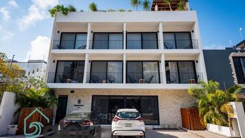 A modern multi-story hotel with a white facade, balconies, and a parking area with two cars.