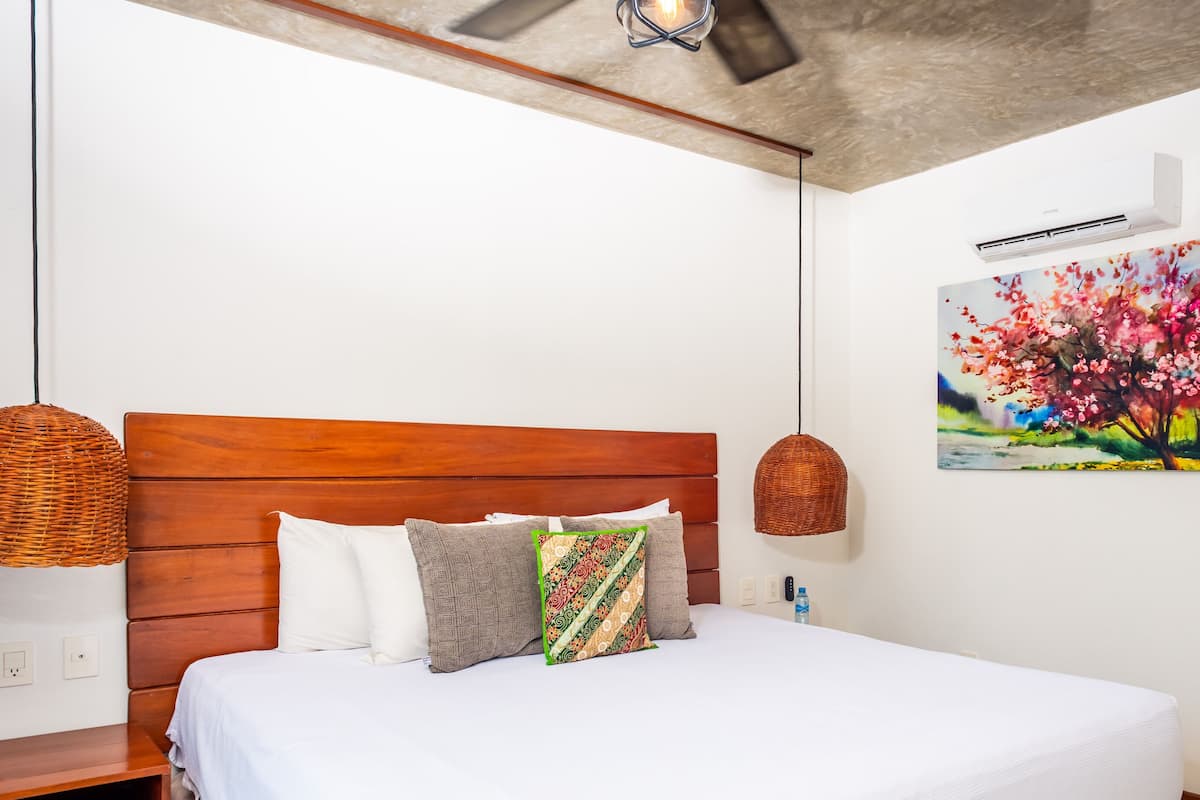 A bedroom with a wooden headboard, a bed with white bedding, patterned pillows, a wicker lamp, and a painting of flowers on the wall.